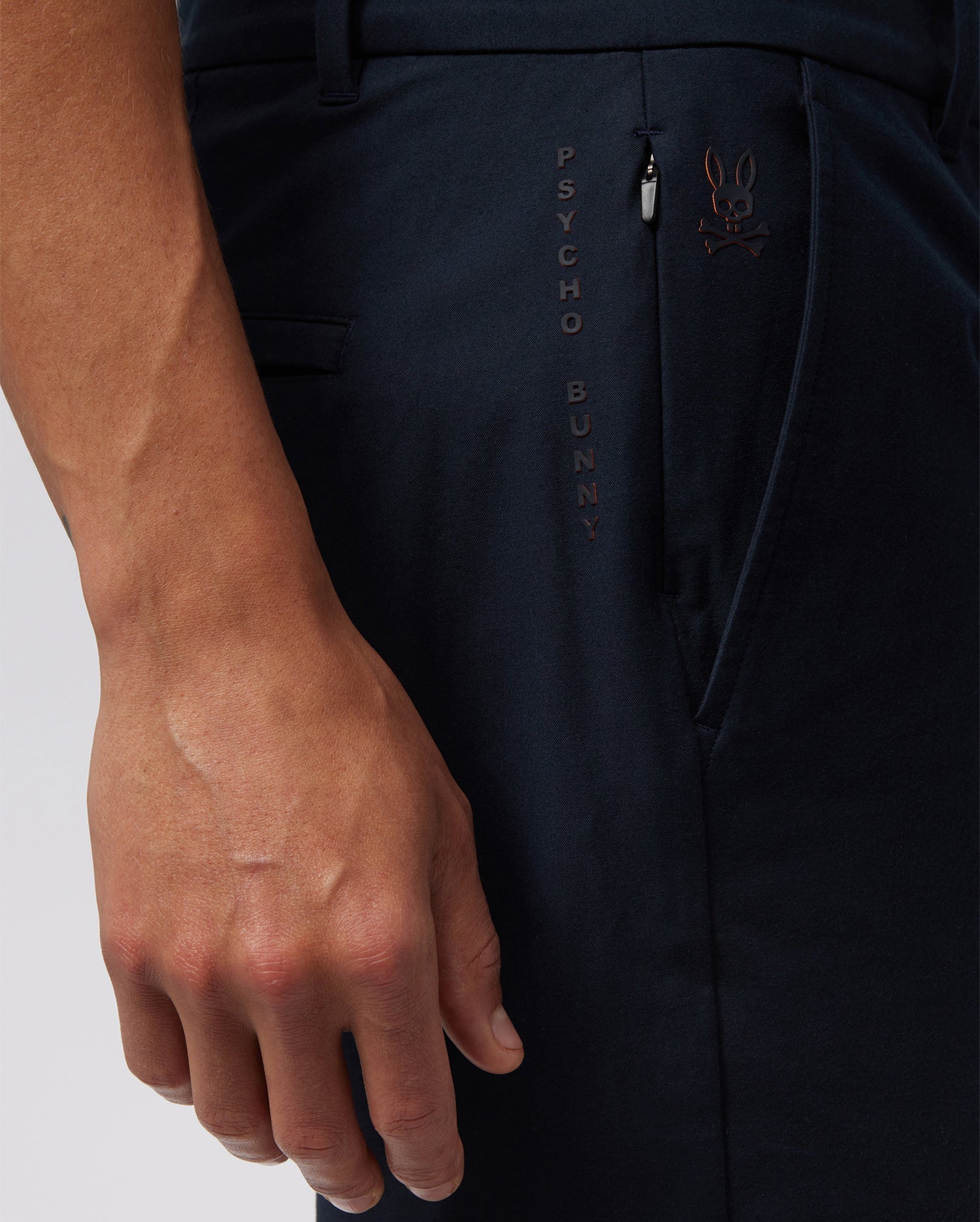 Close-up of a person wearing the MENS GABLE REGULAR FIT SPORT SHORT - B6R761ARCN by Psycho Bunny in dark-colored durable stretch cotton-nylon with the words "PSYCHO BUNNY" embroidered vertically next to the hidden zip pocket. The person's hand is relaxed by their side, partially revealing the right arm and a small logo of a bunny with crossbones.