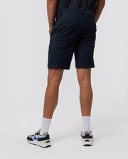 A person standing with their back to the camera, wearing Psycho Bunny MENS GABLE REGULAR FIT SPORT SHORT - B6R761ARCN made from durable stretch cotton-nylon, white socks, and white sneakers with blue and black accents. The background is a plain light gray.