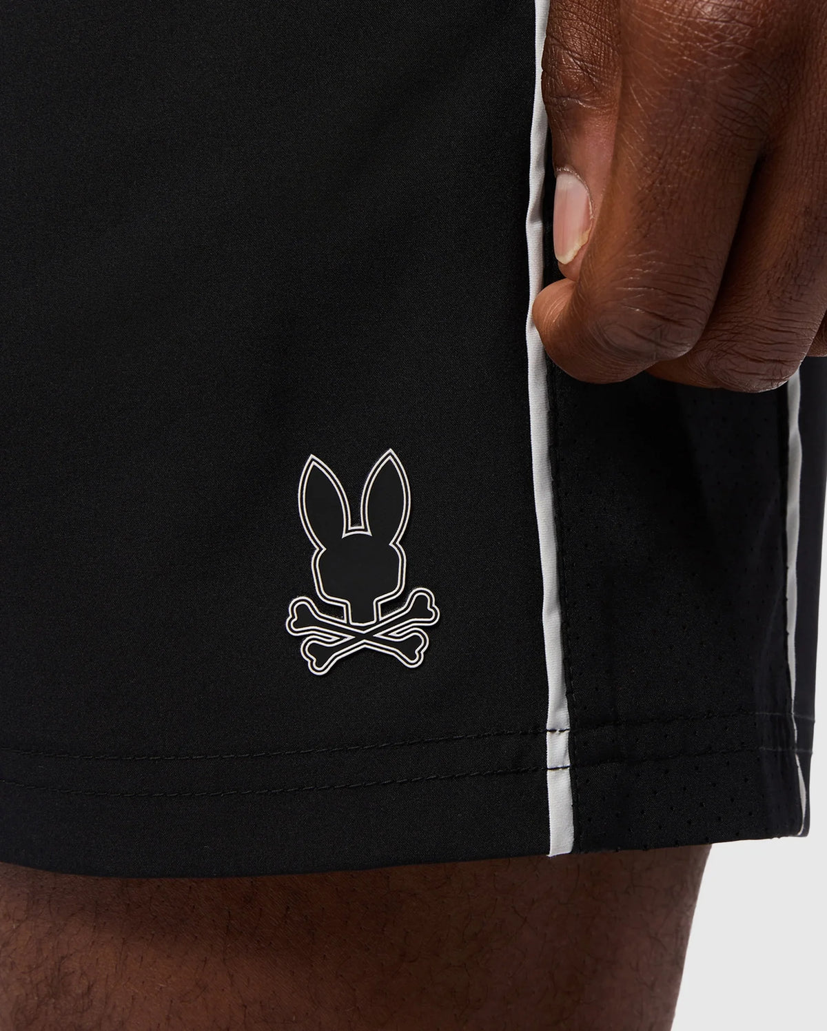 A close-up of a hand resting on the side of Psycho Bunny's MENS MANSFIELD SPORT SHORT - B6R807D200, crafted from breathable polyester. These regular fit black shorts showcase a small bunny head and crossbones logo along with white vertical stripes running down the side seam.