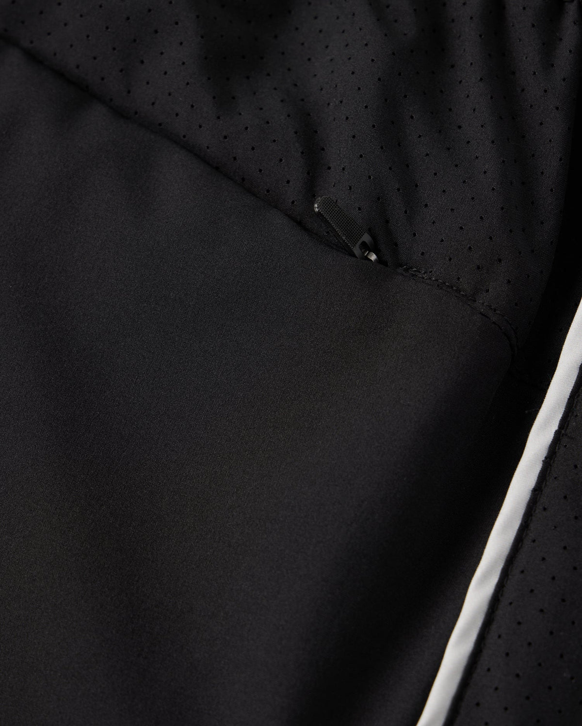 Close-up of a black polyester garment designed by Psycho Bunny, showcasing a subtle, perforated pattern and a partially visible zipper. The fabric exhibits a smooth texture with a hint of shine, highlighted by the MENS MANSFIELD SPORT SHORT style - B6R807D200. It features Psycho Bunny's signature regular fit adorned with a white stripe along the lower edge.