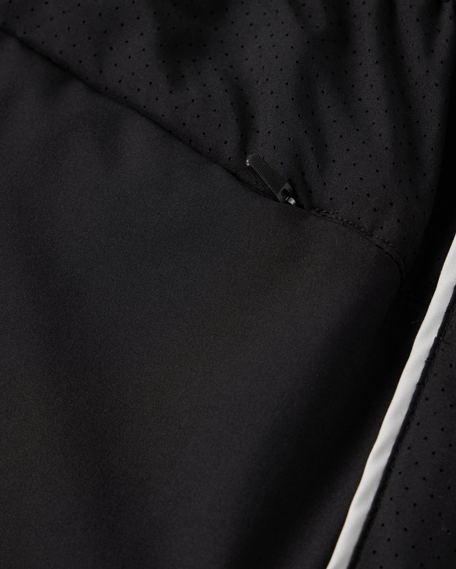 Close-up of a black polyester garment designed by Psycho Bunny, showcasing a subtle, perforated pattern and a partially visible zipper. The fabric exhibits a smooth texture with a hint of shine, highlighted by the MENS MANSFIELD SPORT SHORT style - B6R807D200. It features Psycho Bunny's signature regular fit adorned with a white stripe along the lower edge.
