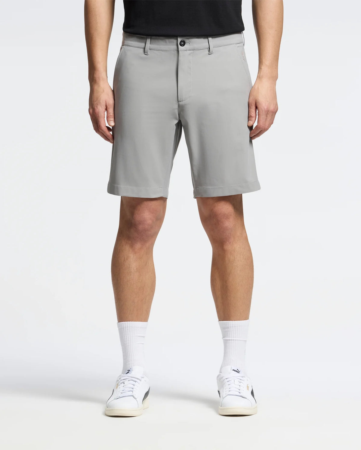 A person wearing Psycho Bunny's MENS 10K STEP SHORT (B6R969F200) stands facing forward in light gray, breathable shorts that reach just above the knees, paired with a black shirt, white sneakers, and white socks against a plain white background.