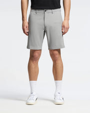 A person wearing Psycho Bunny's MENS 10K STEP SHORT (B6R969F200) stands facing forward in light gray, breathable shorts that reach just above the knees, paired with a black shirt, white sneakers, and white socks against a plain white background.