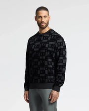 A bearded man with short hair is dressed in a fashionable black Psycho Bunny MENS ROMEO LIGHTWEIGHT SWEATSHIRT - B6S129E200 featuring a patterned design, paired with gray pants. He stands against a plain white background, looking forward with a neutral expression.