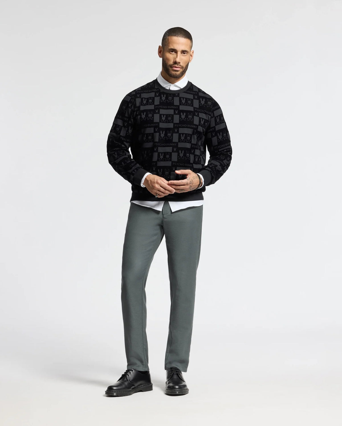 A man stands against a plain background, wearing a stylish black MENS ROMEO LIGHTWEIGHT SWEATSHIRT by Psycho Bunny, paired with a white collared shirt, gray pants, and black shoes. He is posing with his hands together and looking directly at the camera.