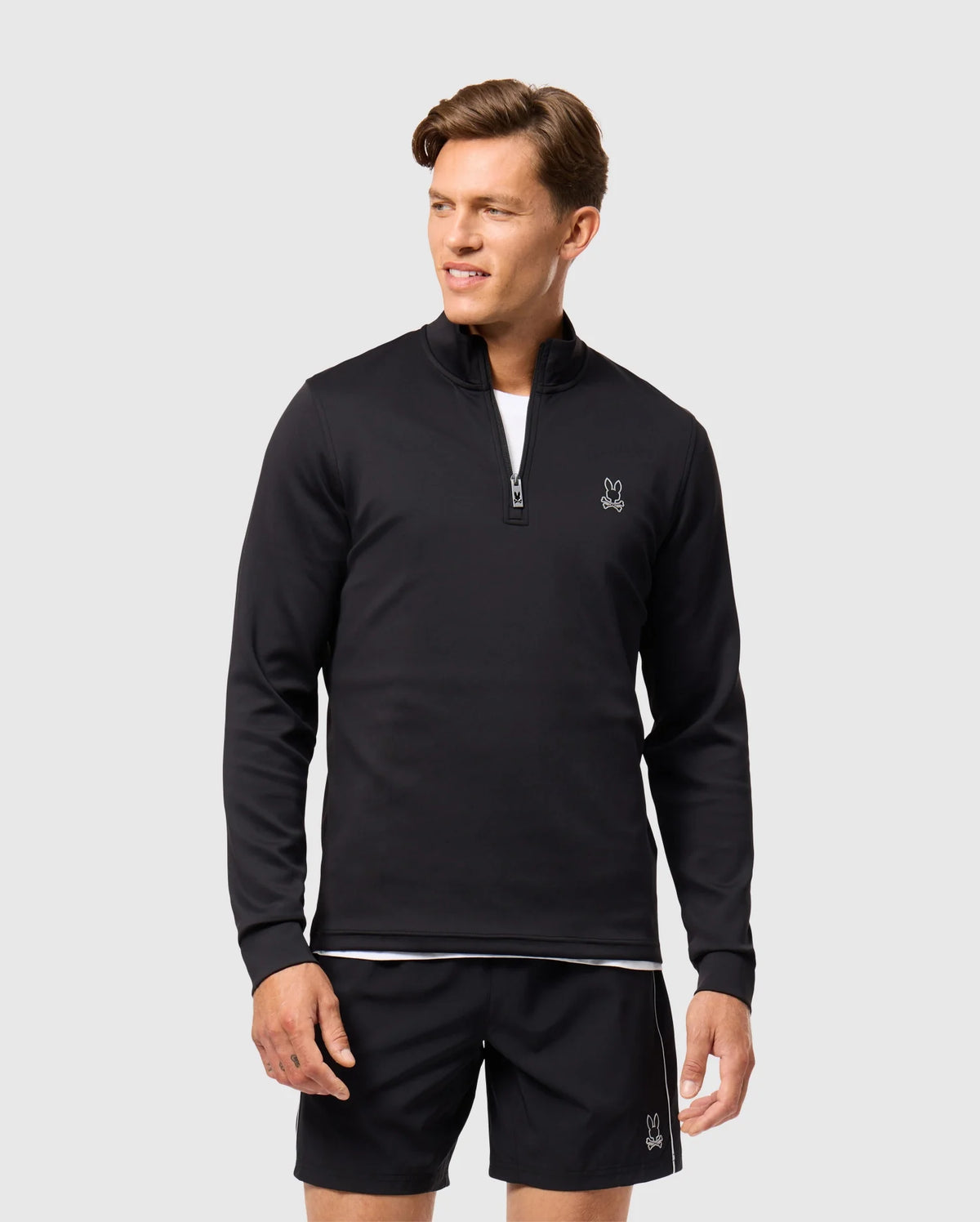 A man wearing a black Psycho Bunny Laurier 1/4 zip hoodie and black shorts stands against a plain white background. The cotton jersey hoodie, featuring a small embroidered logo on the chest, complements his athleisure aesthetic. He looks to the side with a relaxed expression.