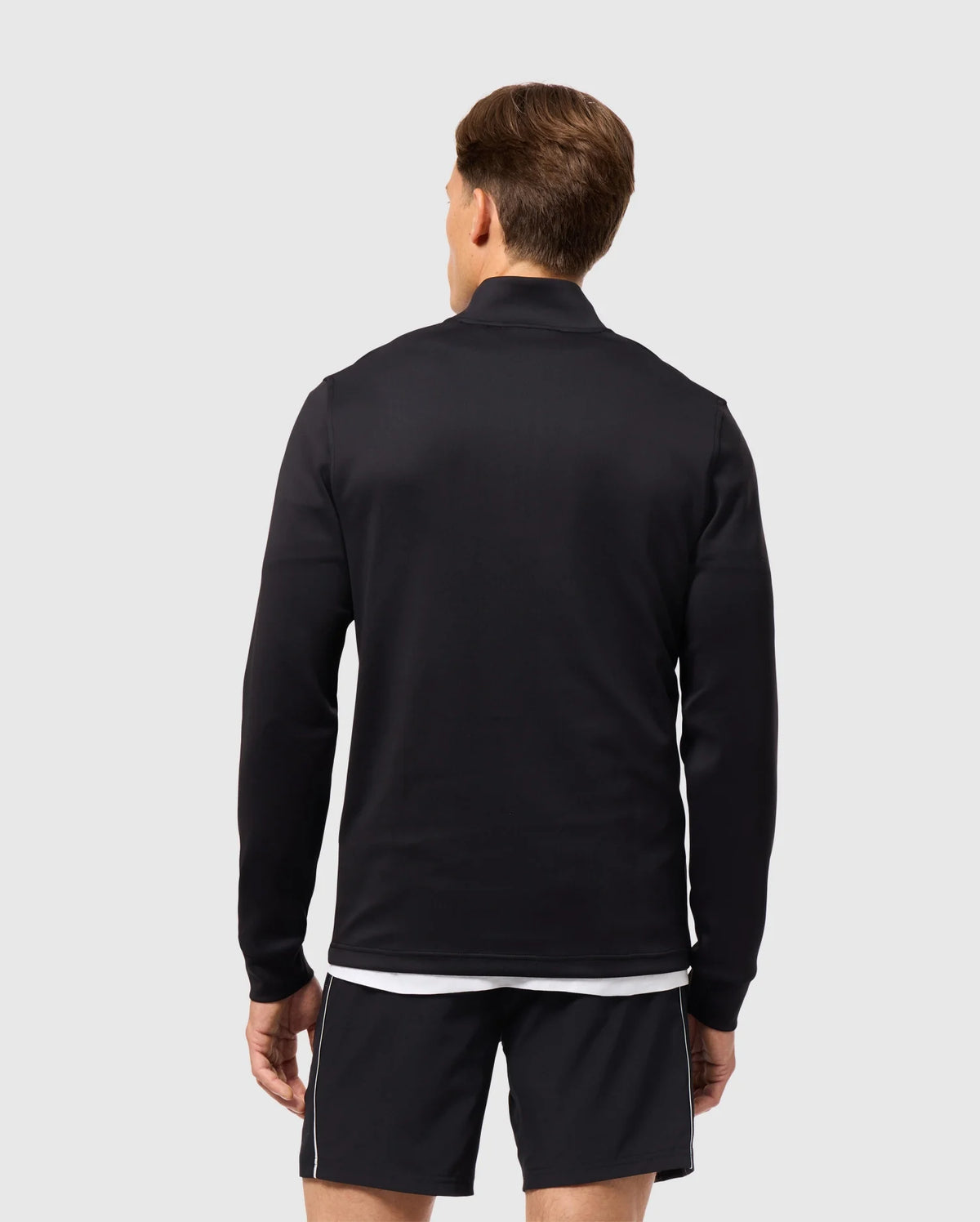 A person stands with their back to the camera, exuding an athleisure vibe in a black MENS LAURIER 1/4 ZIP HOODIE by Psycho Bunny, paired with black shorts featuring white trim, set against a plain light gray background.