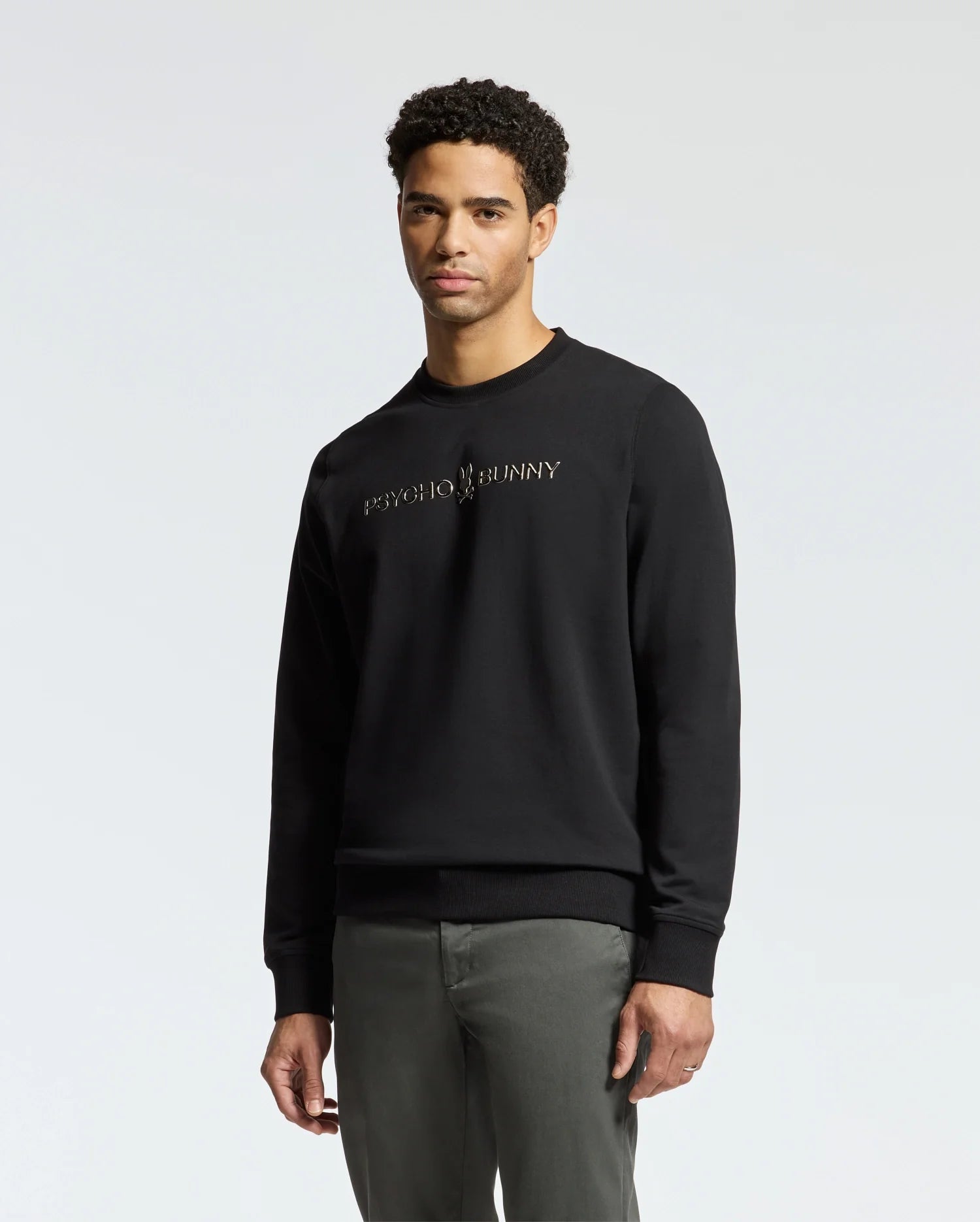 A man wearing the stylish MENS LAVETA FRENCH TERRY SWEATSHIRT - B6S159E200 by Psycho Bunny stands against a plain white background. Crafted from comfortable cotton, this trendy black crew sweatshirt pairs well with his gray pants and complements his short, curly hair.