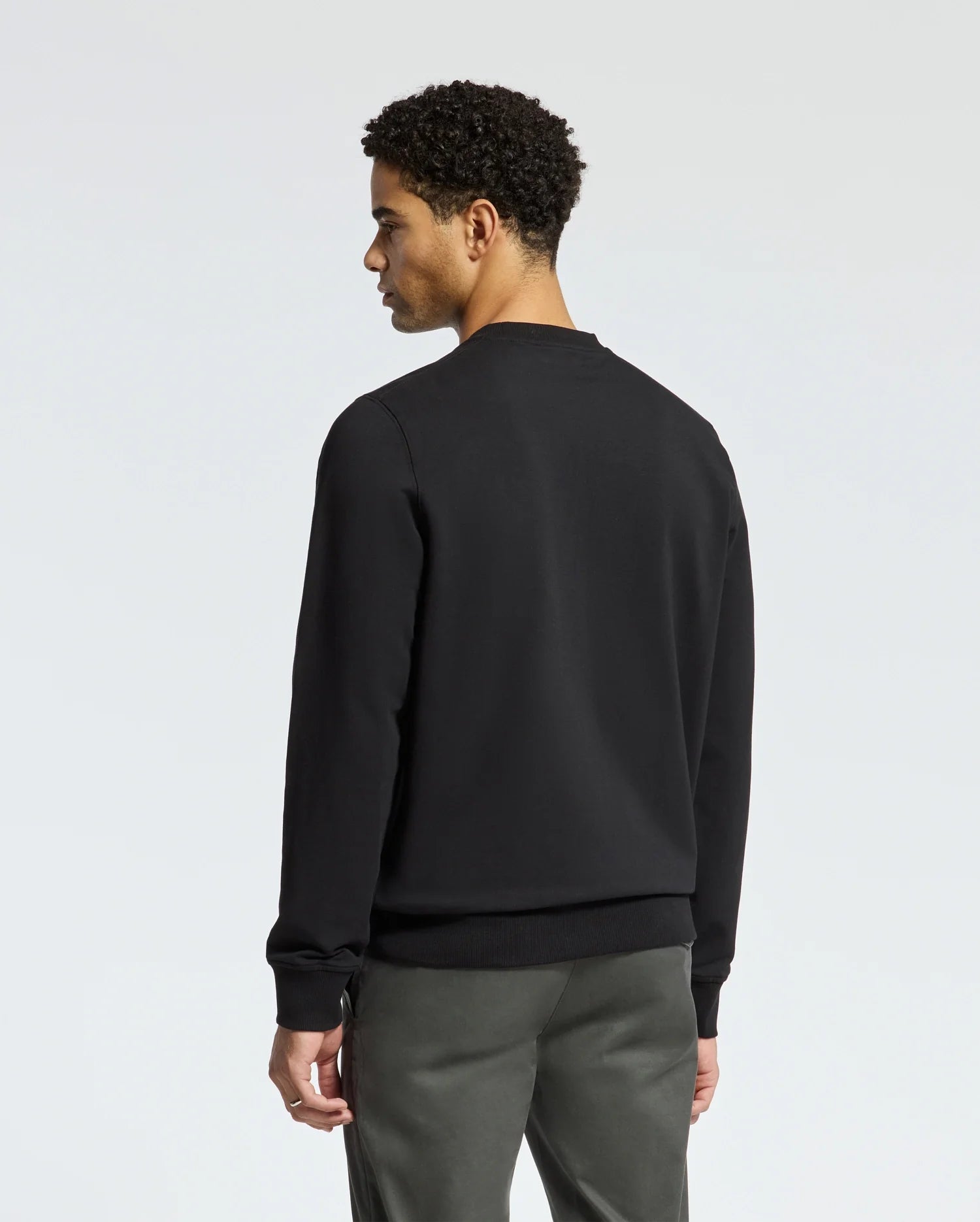 A person with short curly hair stands facing away from the camera, wearing a trendy Psycho Bunny MENS LAVETA French Terry Sweatshirt - B6S159E200 in black and gray pants against a plain light gray background.