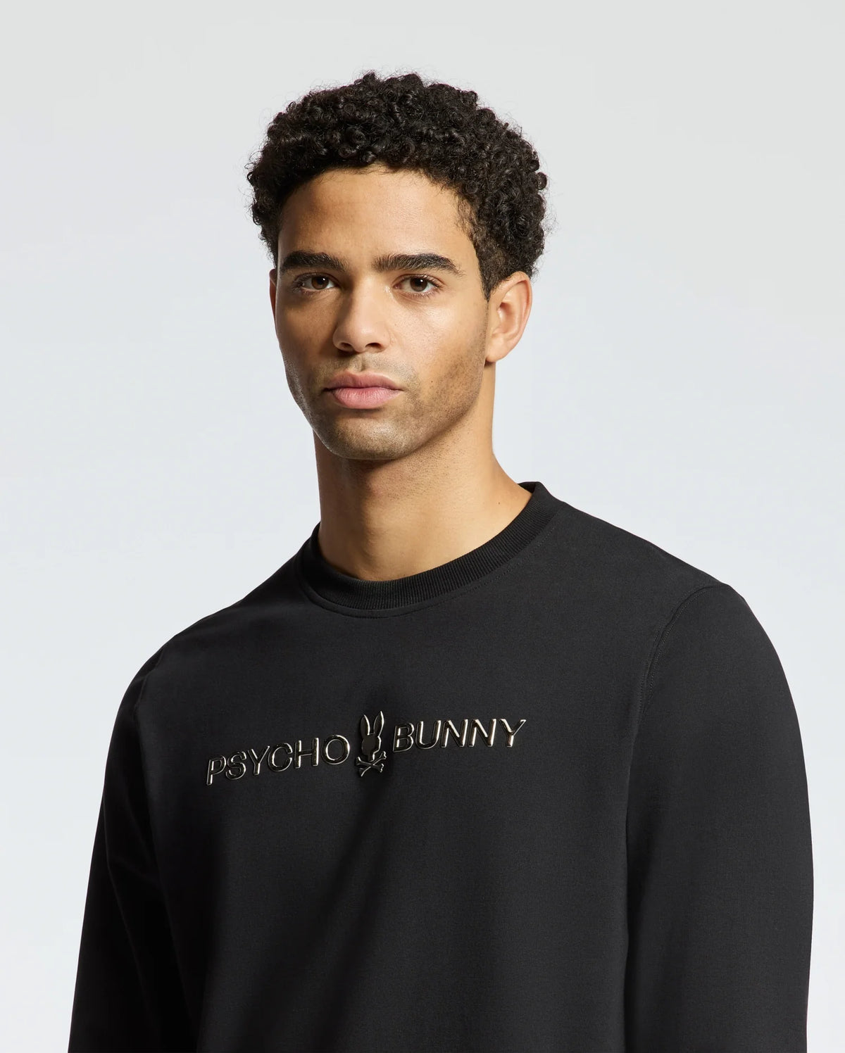 A person wearing the stylish black MENS LAVETA FRENCH TERRY SWEATSHIRT - B6S159E200 from Psycho Bunny stands against a plain, light background. This cozy cotton crew sweatshirt showcases the brand logo and name on the front. The individual has short curly hair and a neutral expression.