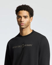 A person wearing the stylish black MENS LAVETA FRENCH TERRY SWEATSHIRT - B6S159E200 from Psycho Bunny stands against a plain, light background. This cozy cotton crew sweatshirt showcases the brand logo and name on the front. The individual has short curly hair and a neutral expression.