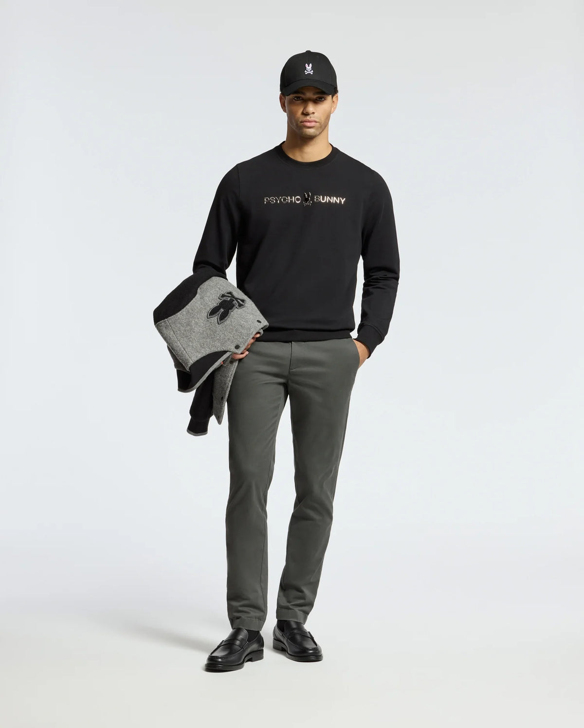 A person poses against a plain background, sporting the MENS LAVETA FRENCH TERRY SWEATSHIRT - B6S159E200 by Psycho Bunny in black, along with gray pants and black shoes. They hold a gray and black jacket and wear a black cap featuring the distinctive bunny logo.