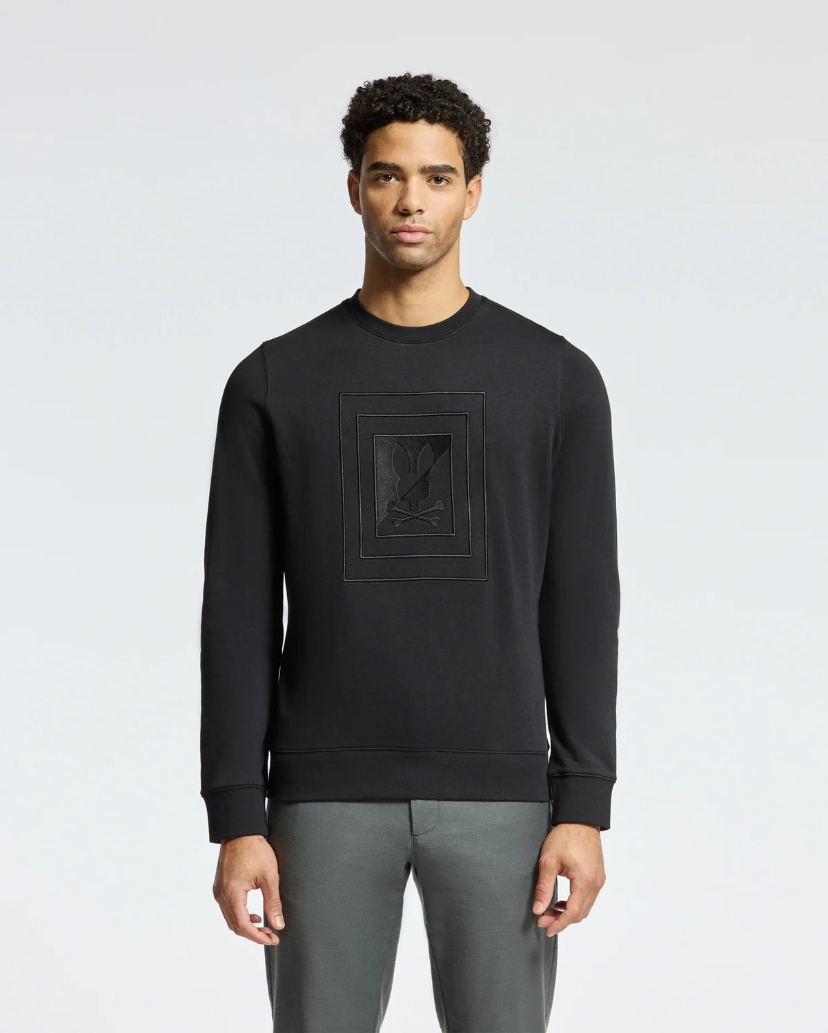 A person wearing the Psycho Bunny MENS OAK CITY CREW NECK - B6S274E200 sweatshirt, featuring raised embroidery and paired with gray pants, stands against a plain light background. The lightweight crew neck design and premium quality fabric enhance the subtle yet stylish appearance.