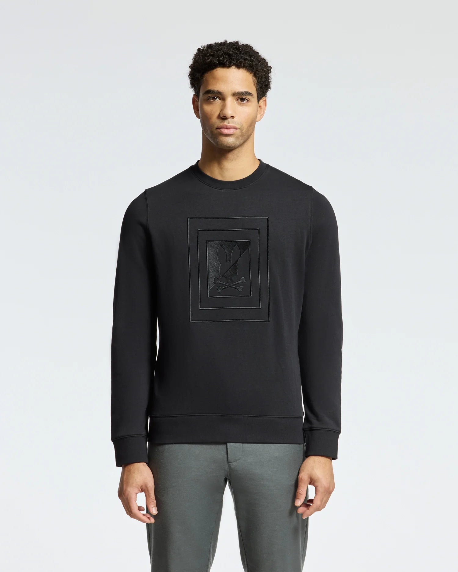 A person wearing the Psycho Bunny MENS OAK CITY CREW NECK - B6S274E200 sweatshirt, featuring raised embroidery and paired with gray pants, stands against a plain light background. The lightweight crew neck design and premium quality fabric enhance the subtle yet stylish appearance.