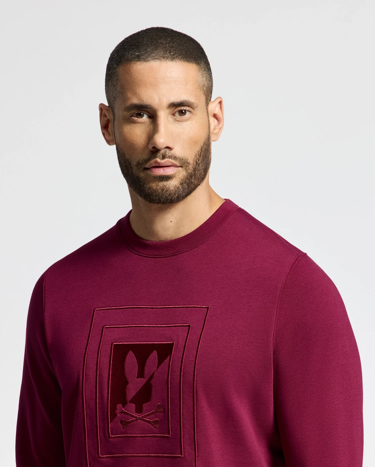 A man with a buzz cut and beard is dressed in a Psycho Bunny MENS OAK CITY CREW NECK - B6S274E200 sweater, which features premium quality maroon fabric and raised embroidery on the front. The background is plain white.