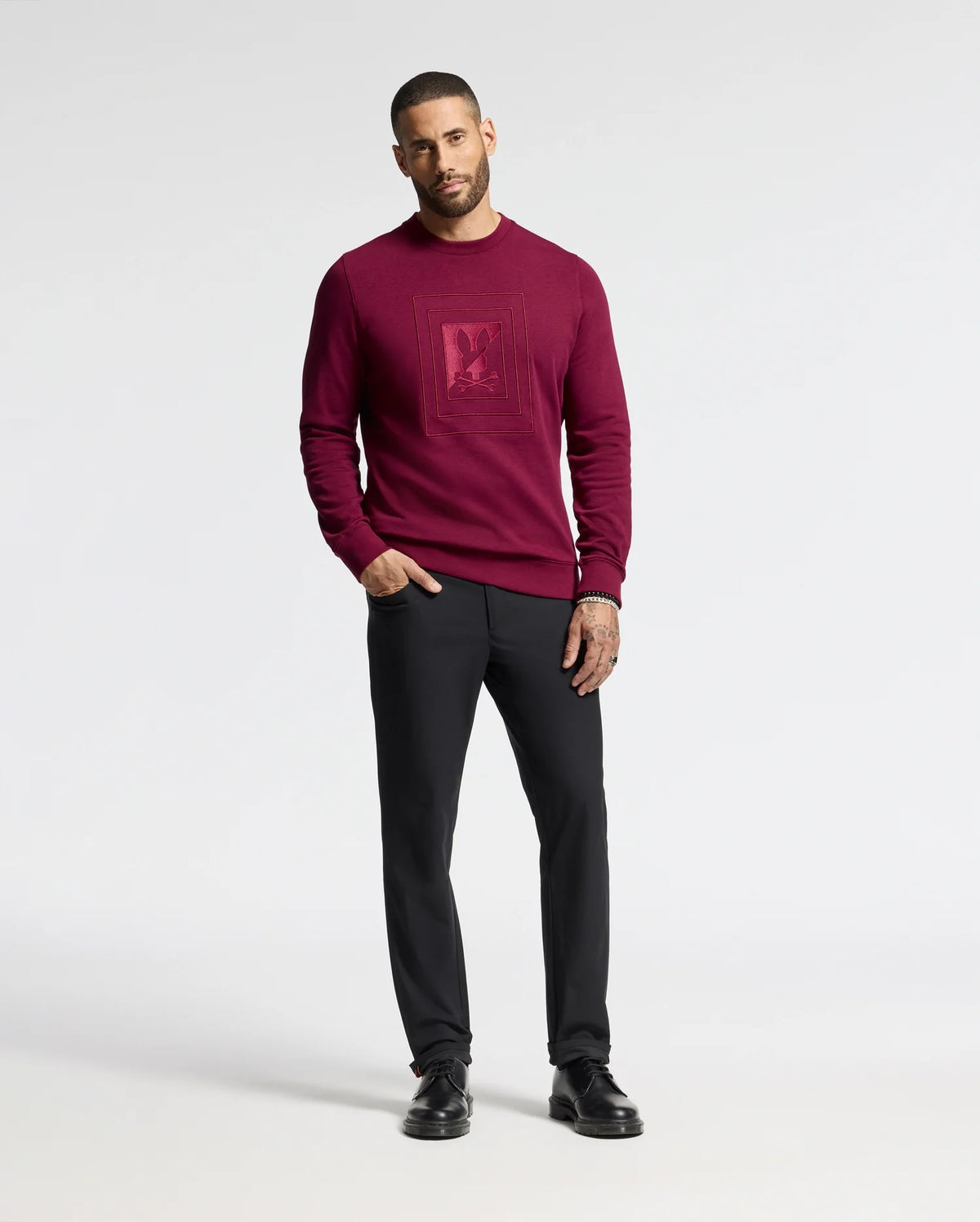A person is positioned against a simple backdrop, dressed in the MENS OAK CITY CREW NECK - B6S274E200 sweater from Psycho Bunny. The sweater is dark red and features raised embroidery. They are paired with black pants and black shoes. The individual has a short haircut and a beard, with their left hand casually placed in their pocket.