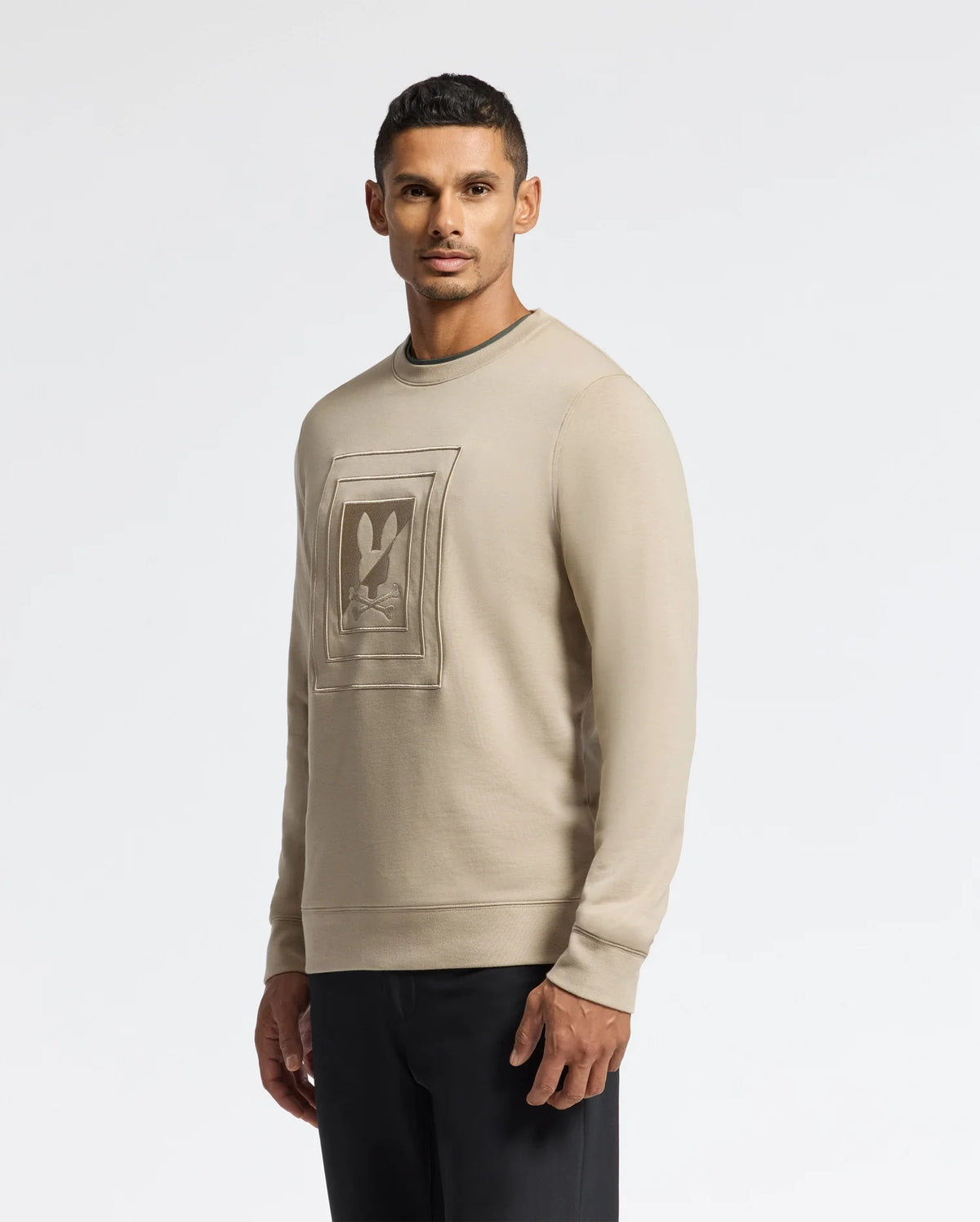 A man wearing the Psycho Bunny MENS OAK CITY CREW NECK - B6S274E200, a premium quality beige sweatshirt with raised embroidery, stands against a plain white background. He is looking directly at the camera, showcasing his short black hair and dark pants.