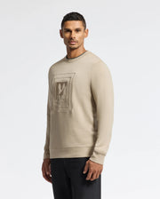A man wearing the Psycho Bunny MENS OAK CITY CREW NECK - B6S274E200, a premium quality beige sweatshirt with raised embroidery, stands against a plain white background. He is looking directly at the camera, showcasing his short black hair and dark pants.