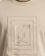 The Psycho Bunny MENS OAK CITY CREW NECK - B6S274E200 is a premium quality beige men's sweater that showcases a raised embroidery of an embossed rectangular design. Within the rectangle, there is a bunny head with crossbones beneath it. The subtle texture highlights contribute to its monochromatic, minimalist appearance.