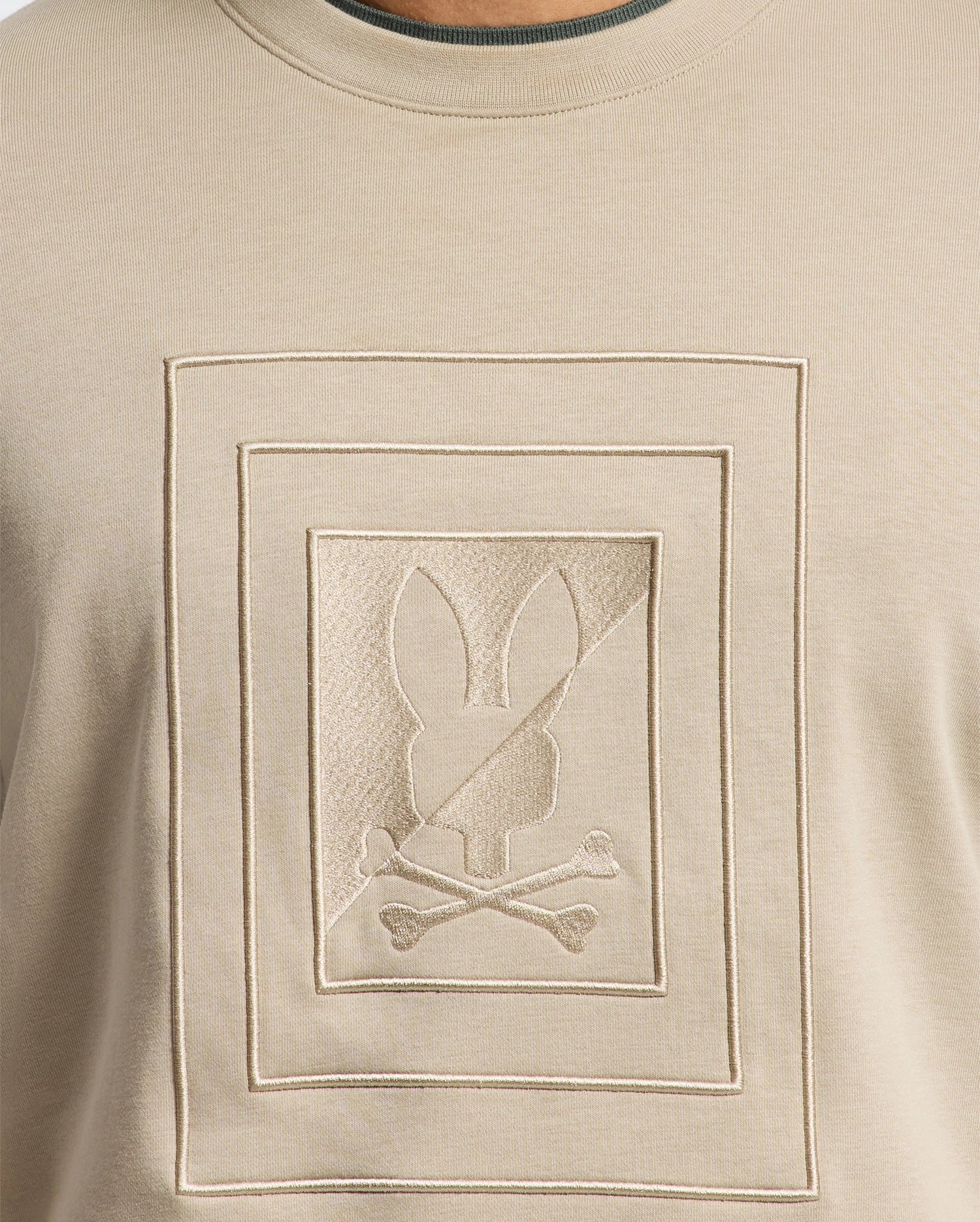 The Psycho Bunny MENS OAK CITY CREW NECK - B6S274E200 is a premium quality beige men's sweater that showcases a raised embroidery of an embossed rectangular design. Within the rectangle, there is a bunny head with crossbones beneath it. The subtle texture highlights contribute to its monochromatic, minimalist appearance.