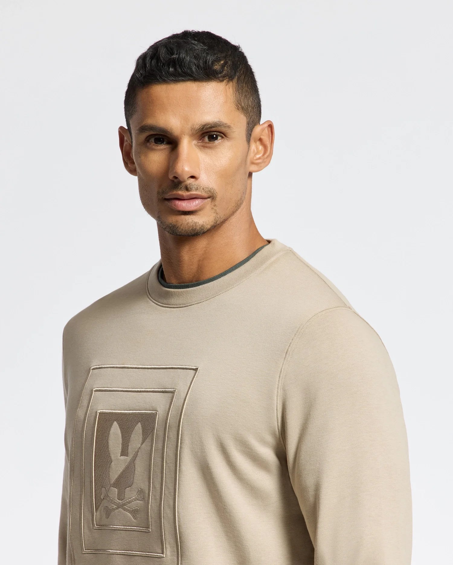 A person with short dark hair is wearing the MENS OAK CITY CREW NECK - B6S274E200 by Psycho Bunny, a beige men's crew neck sweater featuring a rabbit graphic. The premium quality design features raised embroidery as they look directly at the camera against a plain light background.