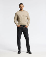 A man stands confidently against a plain background, wearing the Psycho Bunny MENS OAK CITY CREW NECK - B6S274E200, a premium quality beige sweatshirt featuring subtle raised embroidery. He pairs it with black pants and black shoes, his hands in his pockets as he looks straight ahead with a neutral expression.