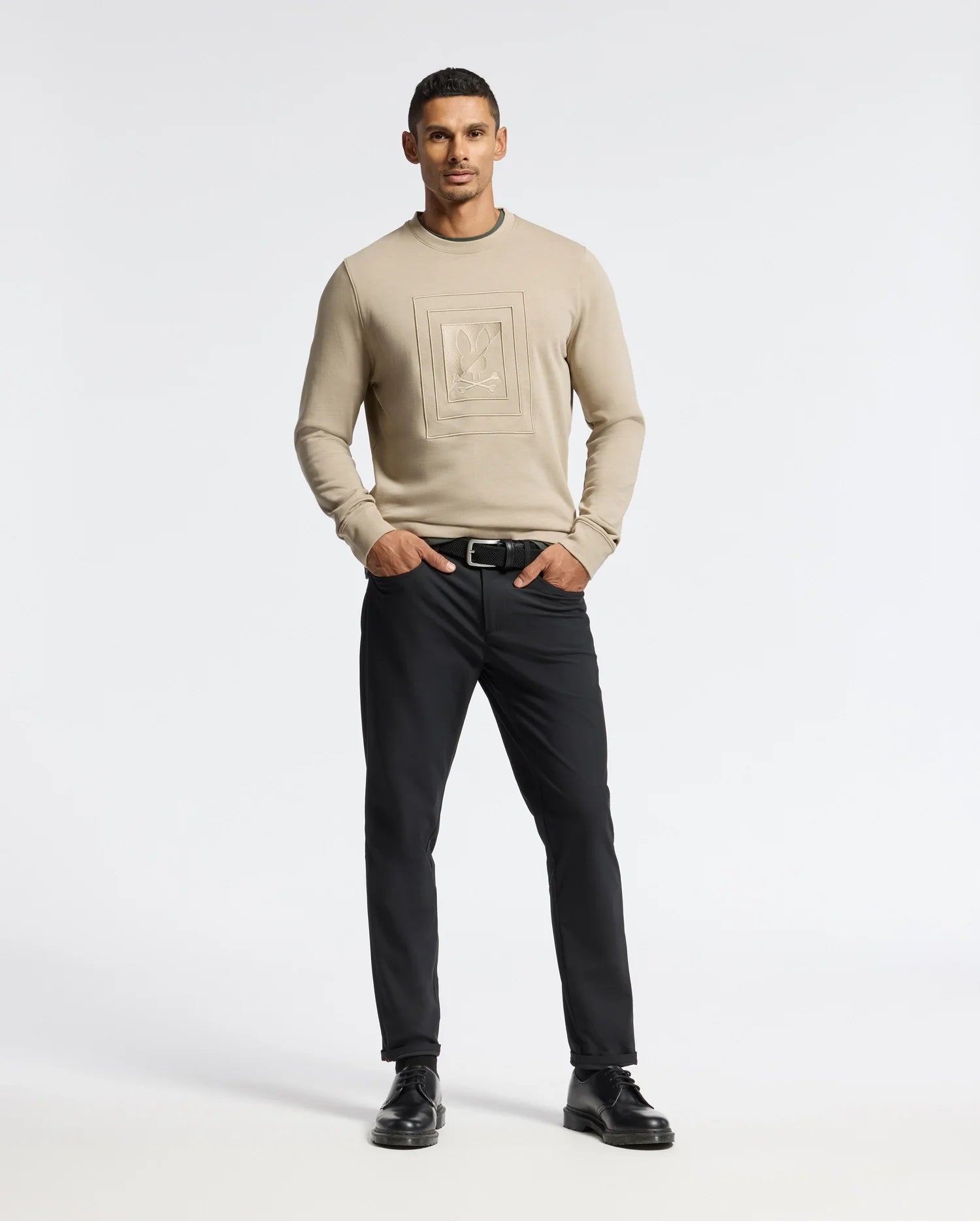 A man stands confidently against a plain background, wearing the Psycho Bunny MENS OAK CITY CREW NECK - B6S274E200, a premium quality beige sweatshirt featuring subtle raised embroidery. He pairs it with black pants and black shoes, his hands in his pockets as he looks straight ahead with a neutral expression.