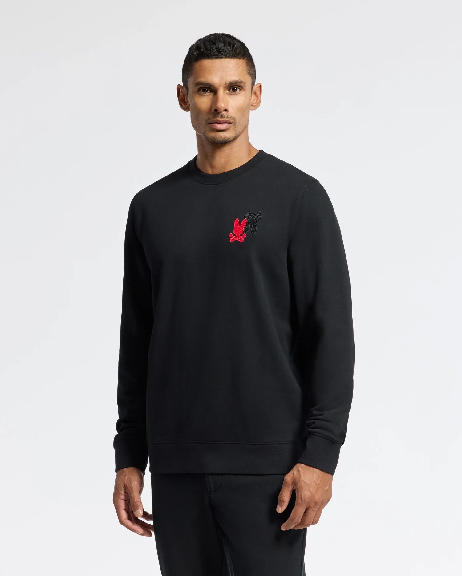 A man wearing the Psycho Bunny MENS PIERCE PEACHED TERRY CREW - B6S278E200, which features an embroidered small red graphic on the chest, stands against a plain background. His hands are relaxed by his sides, and he has short dark hair.