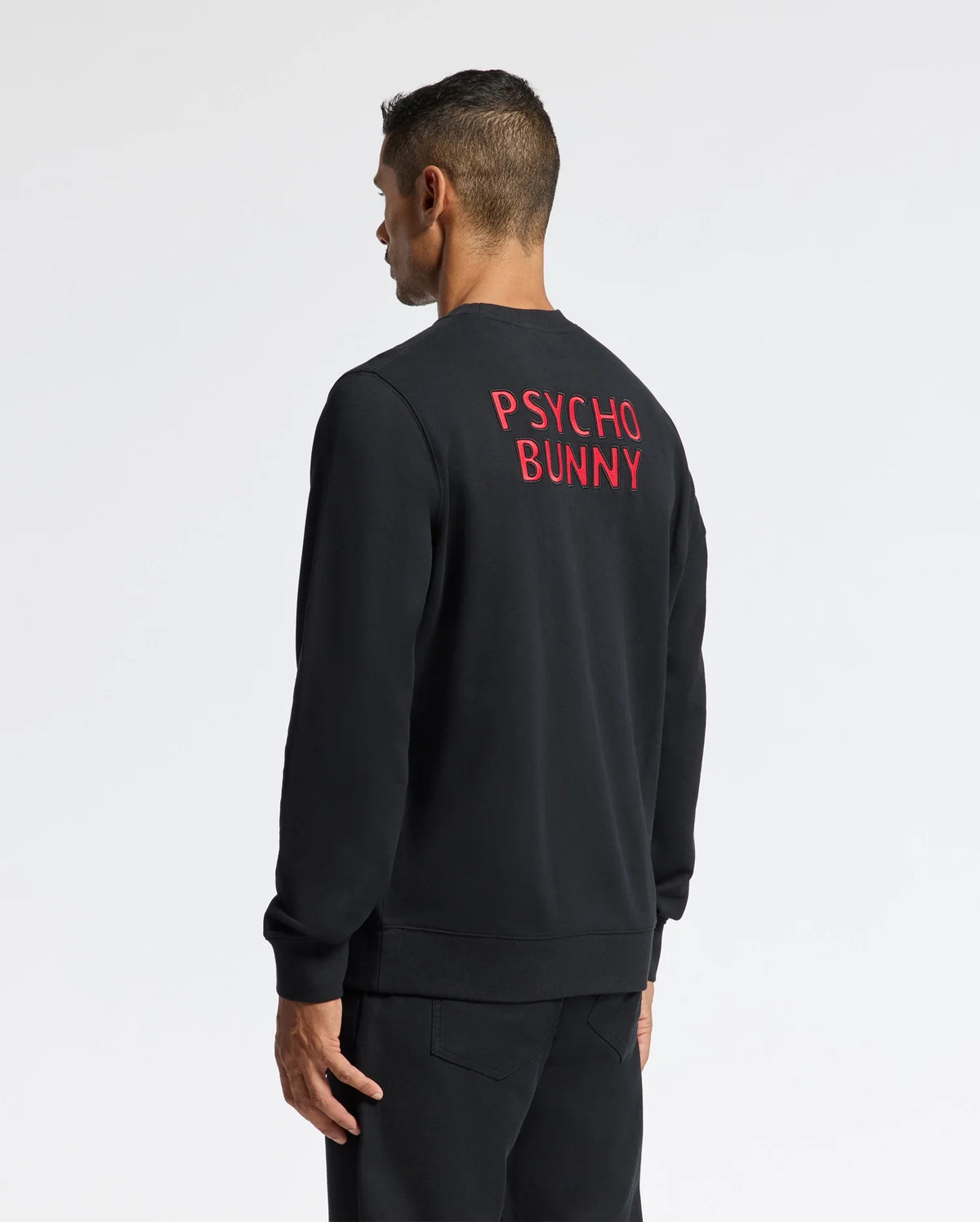 A man is seen wearing the stylish MENS PIERCE PEACHED TERRY CREW - B6S278E200 by Psycho Bunny. This fashionable black sweatshirt features "Psycho Bunny" embroidered in red letters on the back, crafted from high-quality cotton, and stands out against the plain white background as he looks to the side while facing away from the camera.