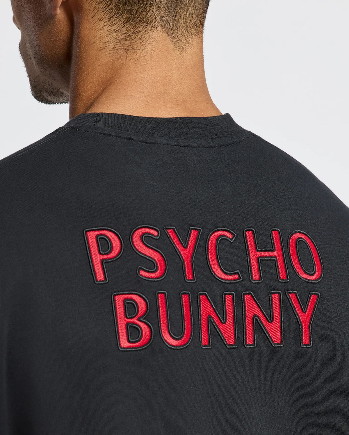 A person is wearing the MENS PIERCE PEACHED TERRY CREW, featuring black embroidered cotton with "PSYCHO BUNNY" in red text on the back. The background is plain white.