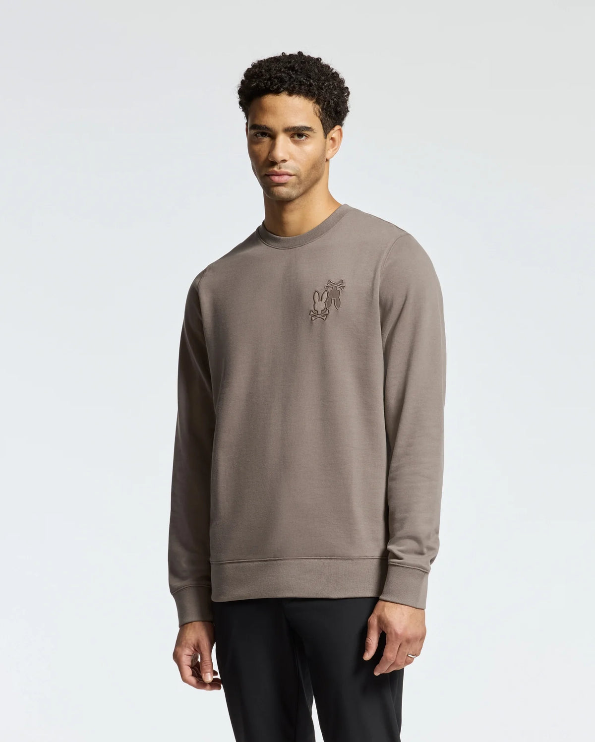 A man in a Psycho Bunny MENS PIERCE PEACHED TERRY CREW - B6S278E200, light brown with chest embroidery, stands against a plain background. Made of high-quality cotton, he has short, curly hair and a neutral expression.