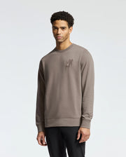 A man in a Psycho Bunny MENS PIERCE PEACHED TERRY CREW - B6S278E200, light brown with chest embroidery, stands against a plain background. Made of high-quality cotton, he has short, curly hair and a neutral expression.