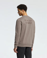 A person with short curly hair is shown from behind, wearing the MENS PIERCE PEACHED TERRY CREW - B6S278E200 by Psycho Bunny. This stylish beige sweatshirt pairs effortlessly with their black pants and features a plain, light-colored background.