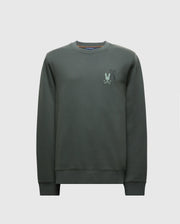 The MENS WORDMARK PEACHED TERRY CREW by Psycho Bunny (B6S278E200) is a dark green pullover made from knit cotton. It features a subtle graphic design on the left chest with a stylized silhouette of a character. With long sleeves and a relaxed fit, this crewneck sweatshirt is ideal for casual style with comfort.
