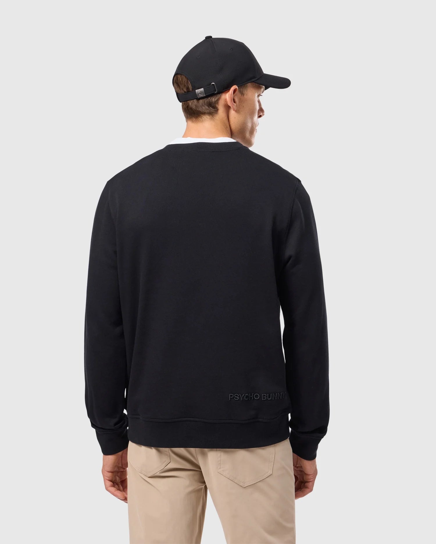 A man wearing the Psycho Bunny MENS KINGSTON SWEATSHIRT - B6S480D200, which features a bold logo with Kingston 3D-embroidery, pairs it with a black cap against a plain gray background. His beige pants highlight the rich cotton jersey material of his striking outfit as he turns away.