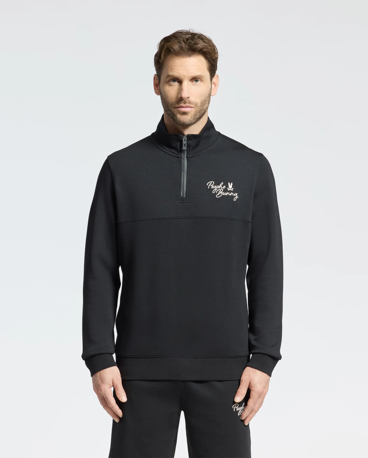 A man wearing the Psycho Bunny MENS DESERT 1/4 MOCK ZIP - B6S488F200 stands against a plain background. The sleek black pullover, featuring an embroidered wordmark logo on the chest, is crafted from 100% cotton for a comfortable and stylish casual outfit.