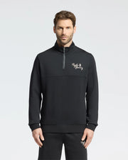A man wearing the Psycho Bunny MENS DESERT 1/4 MOCK ZIP - B6S488F200 stands against a plain background. The sleek black pullover, featuring an embroidered wordmark logo on the chest, is crafted from 100% cotton for a comfortable and stylish casual outfit.