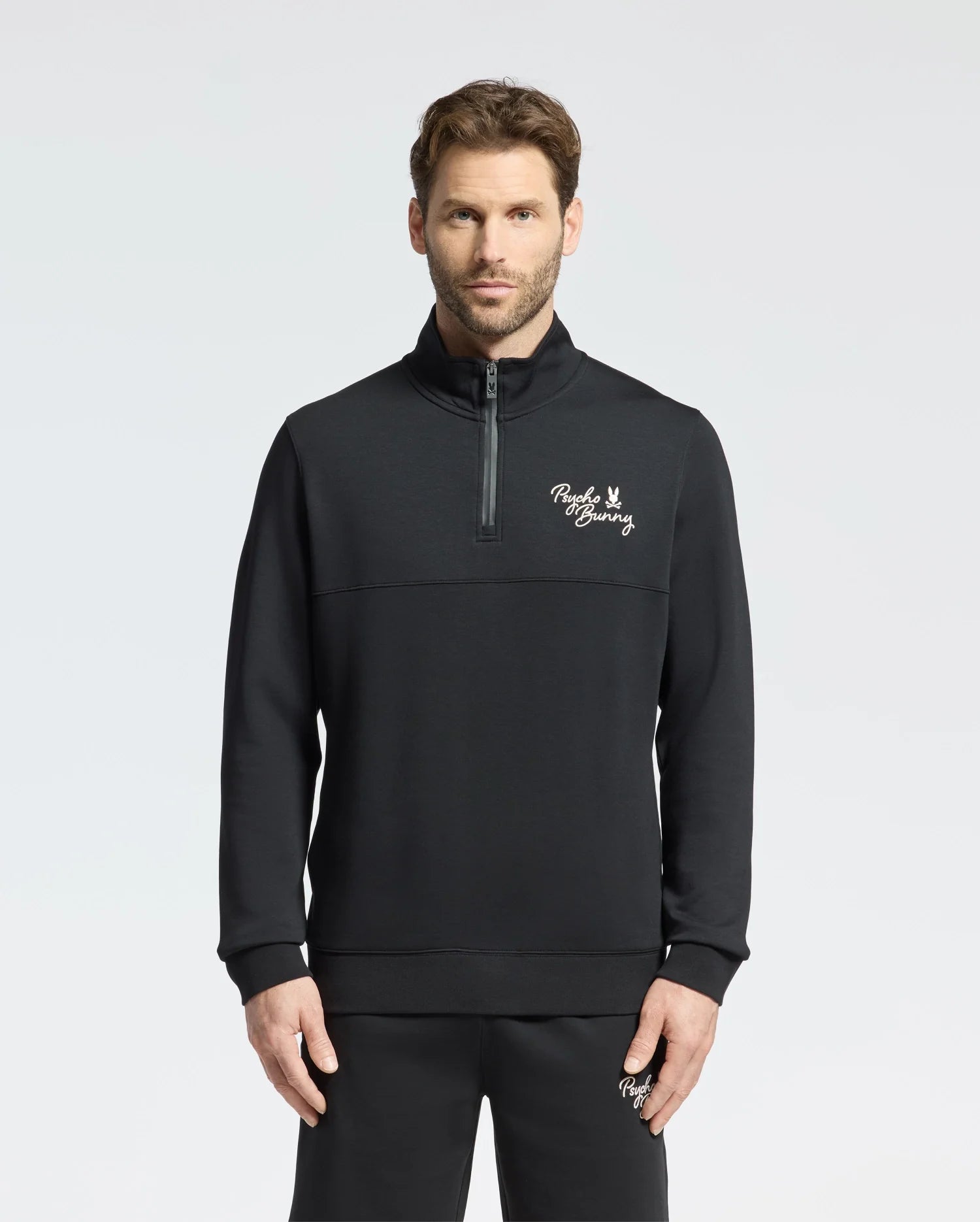 A man wearing the Psycho Bunny MENS DESERT 1/4 MOCK ZIP - B6S488F200 stands against a plain background. The sleek black pullover, featuring an embroidered wordmark logo on the chest, is crafted from 100% cotton for a comfortable and stylish casual outfit.