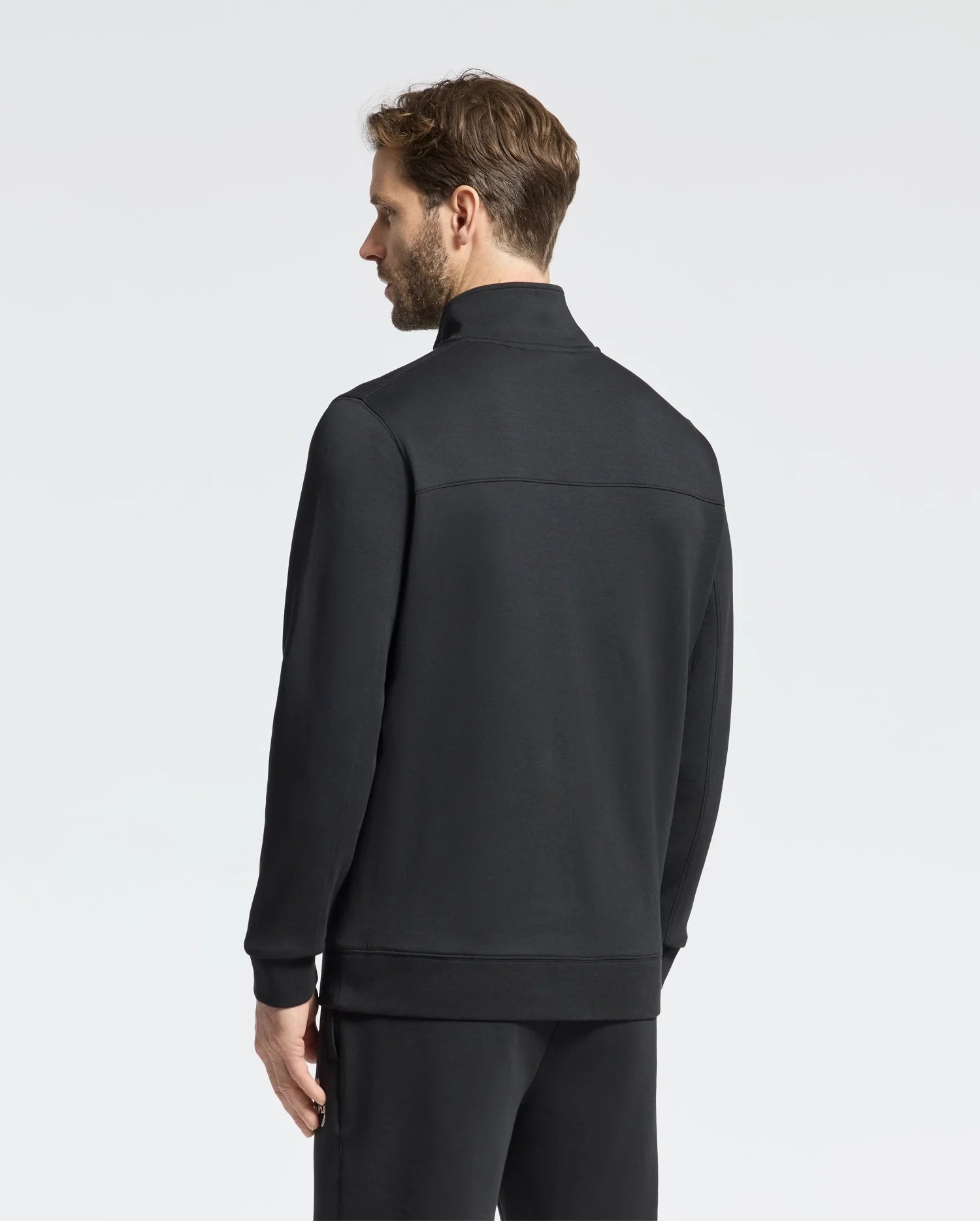 A man with short brown hair stands looking away, donning the Psycho Bunny MENS DESERT 1/4 MOCK ZIP - B6S488F200. This sleek black ensemble features an embroidered wordmark logo and matching pants, with a modern quarter-zip design set against a plain light gray background.