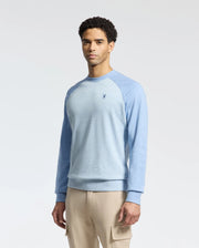 A person is wearing a Psycho Bunny MENS ARCHIE SWEATSHIRT - B6S630F200 in light blue with darker sleeves and beige cargo pants. The embroidered Bunny logo on the chest adds playfulness, while the plain white background enhances the monochrome style.