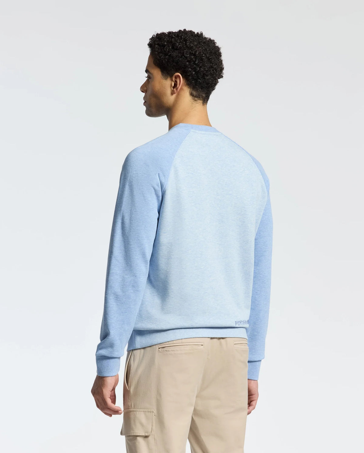With their back to the camera, a person with short curly hair models the MENS ARCHIE SWEATSHIRT by Psycho Bunny, paired with beige pants, suggesting a monochrome look against a plain, light-colored backdrop.