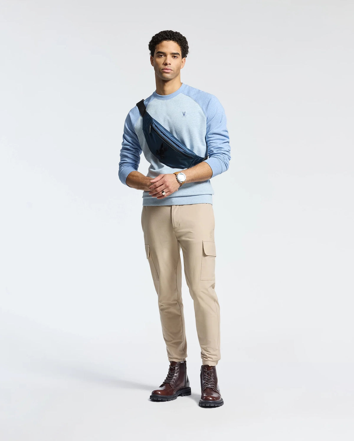 A man in a light blue Psycho Bunny MENS ARCHIE SWEATSHIRT - B6S630F200 and beige cargo pants stands against a plain background. He carries a black crossbody bag, wears brown boots and a watch, with his short, curly hair completing the monochrome look.