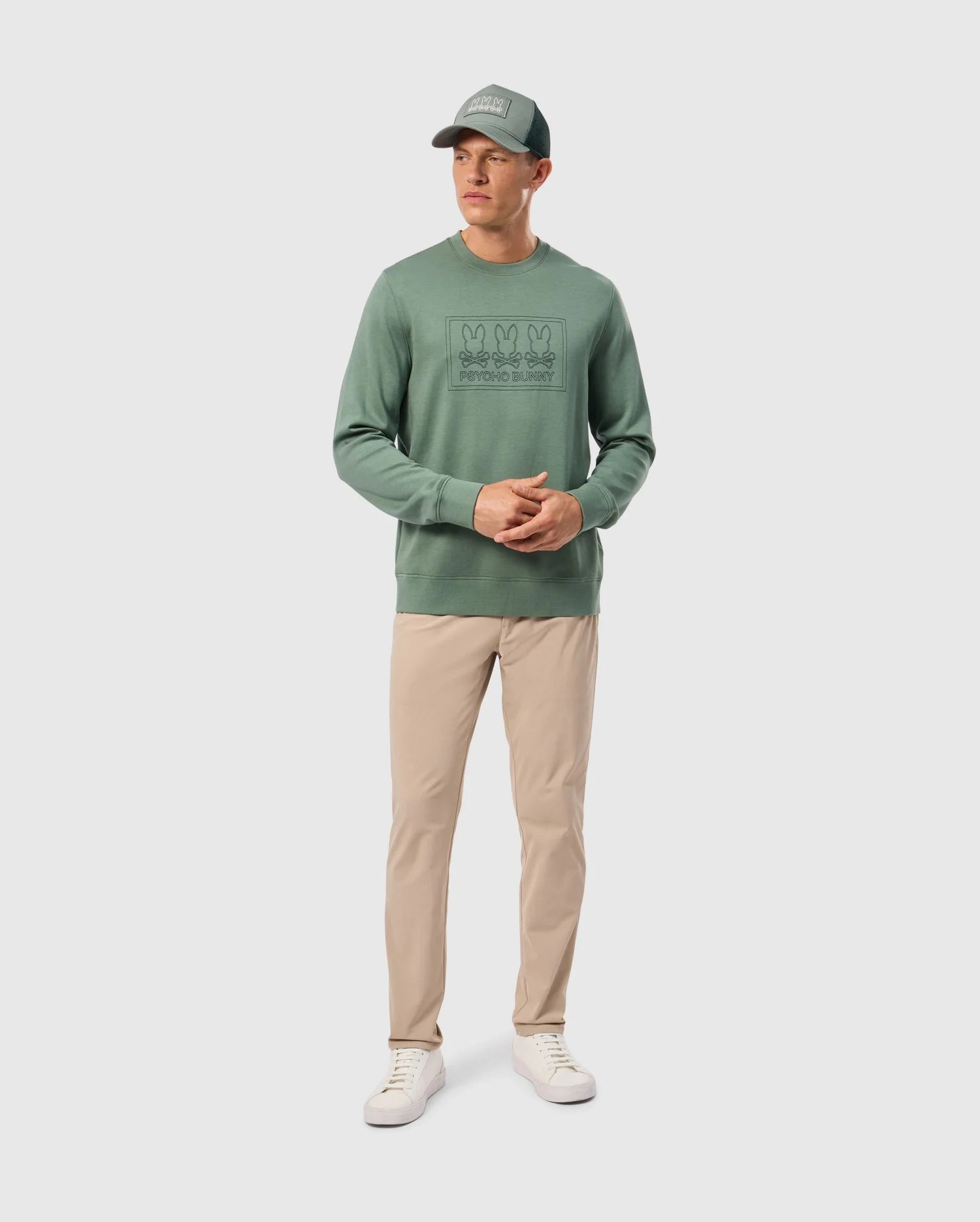 A man wearing the Psycho Bunny MENS TACOMA SWEATSHIRT - B6S927D200 in green, along with beige trousers, white sneakers, and a coordinating green cap stands against a plain backdrop. His outfit, crafted from premium stretch fabric, promises luxurious comfort as he clasps his hands together in front of him.