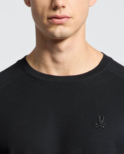 A person wears a Psycho Bunny black Pima cotton piqué long-sleeve tee, featuring an embroidered Bunny with crossed bones on the chest. The background is plain and light-colored, highlighting the upper torso and neck area.