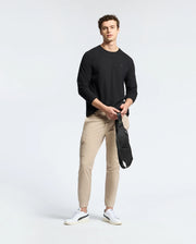 A man stands against a simple background wearing a black Psycho Bunny Men's Taylor Piqué Long Sleeve Tee, beige cargo pants, and white sneakers. He holds an embroidered Bunny crossbody bag in his left hand and looks towards the camera.