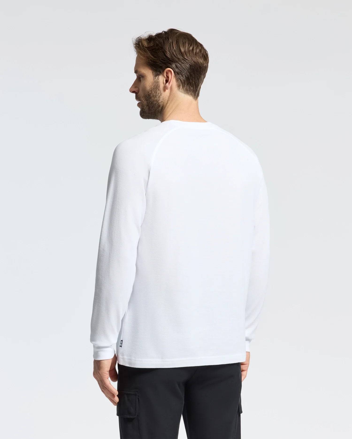 A brown-haired man gazes away from the camera, wearing a crisp white Psycho Bunny MENS TAYLOR PIQUE LONG SLEEVE TEE with raglan sleeves and black pants, against a plain background.