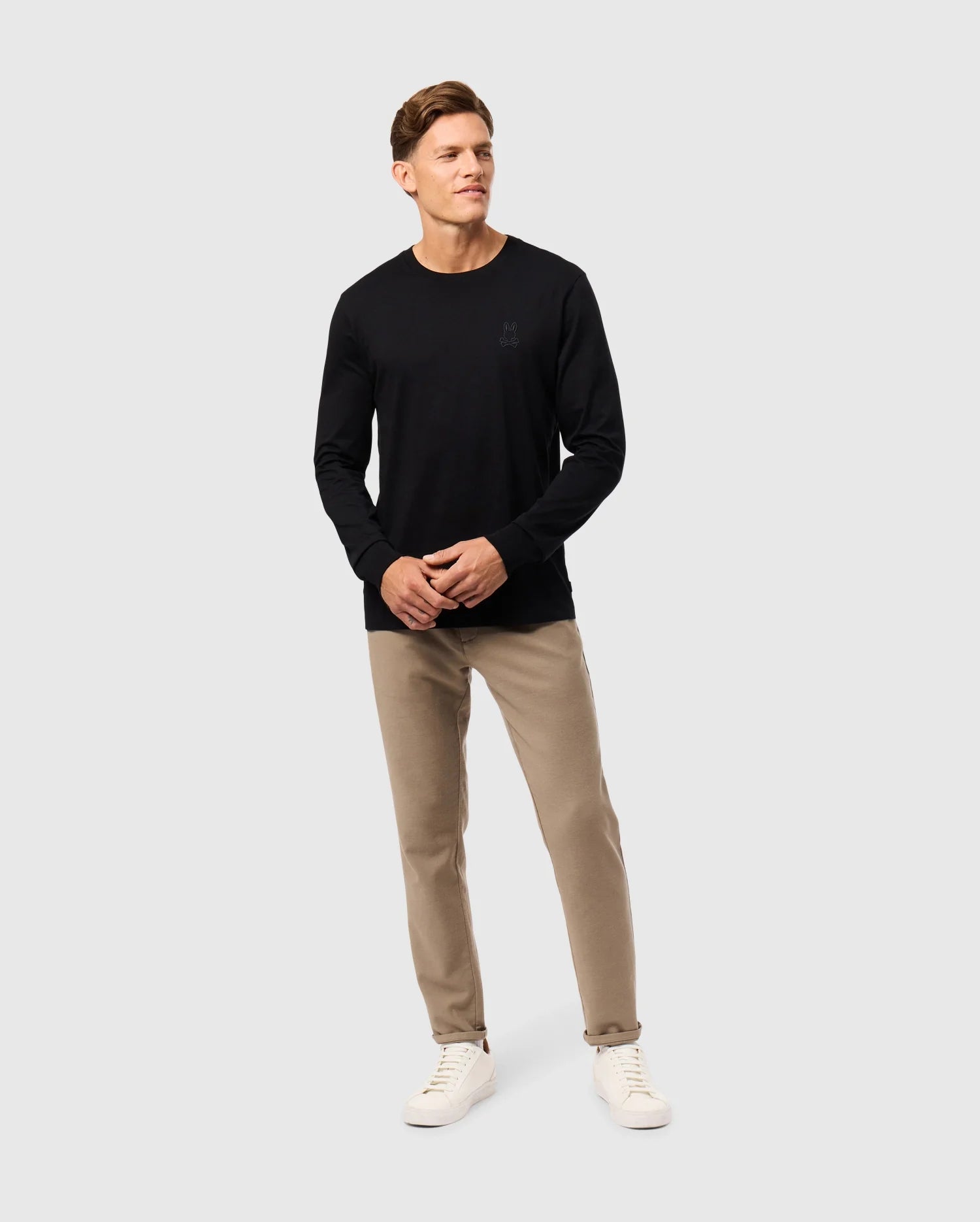 A person stands against a plain white background wearing a Psycho Bunny MENS ALBERTON LONG SLEEVE TEE in black, made of Pima cotton, and beige pants. They are also sporting white sneakers and have short brown hair, gazing slightly to the side with a relaxed posture—a true fashion masterpiece.