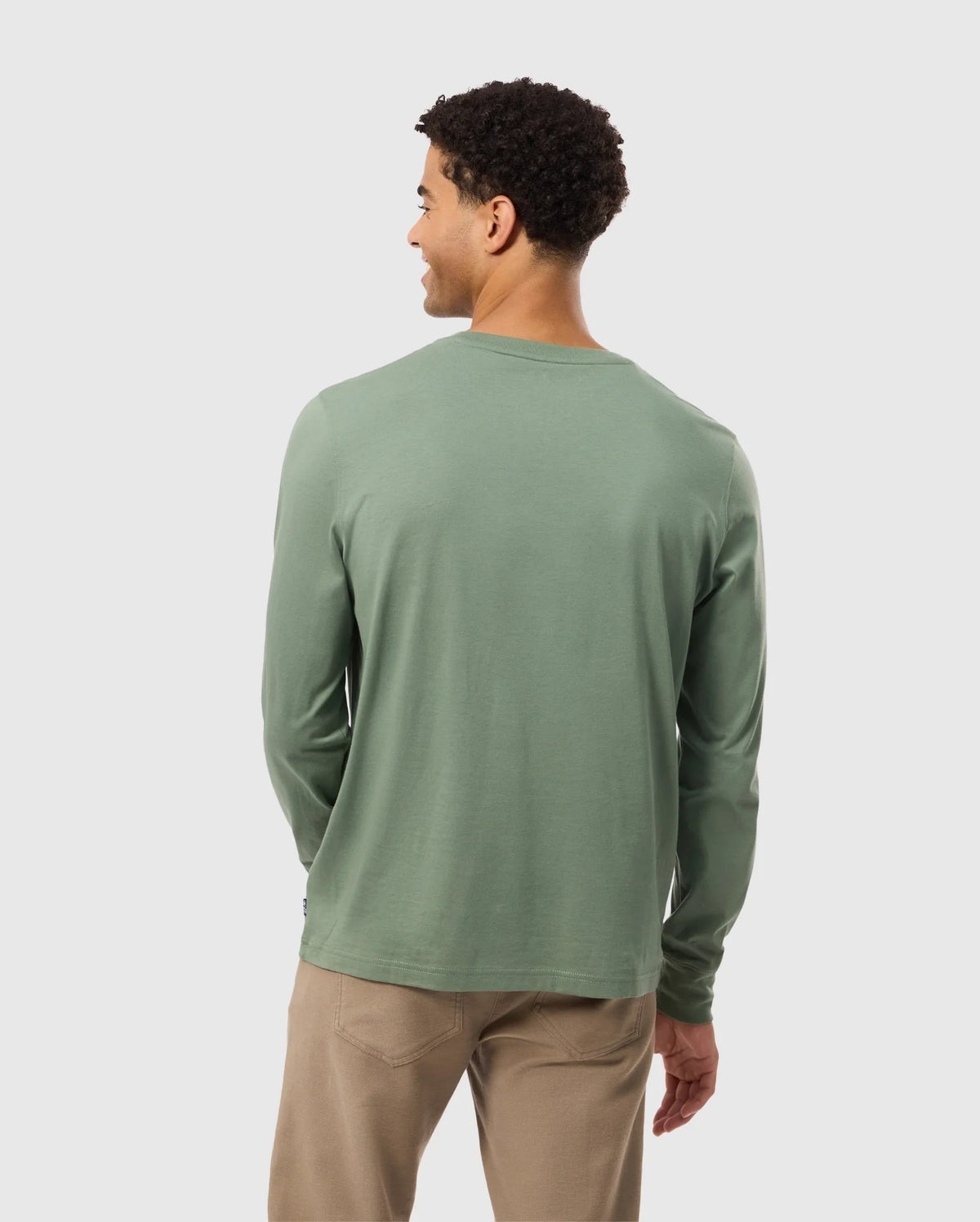 A person with short curly hair is wearing the Psycho Bunny MENS ALBERTON LONG SLEEVE TEE in light green, made from Pima cotton, along with beige pants. They are standing and facing away from the camera against a plain background.
