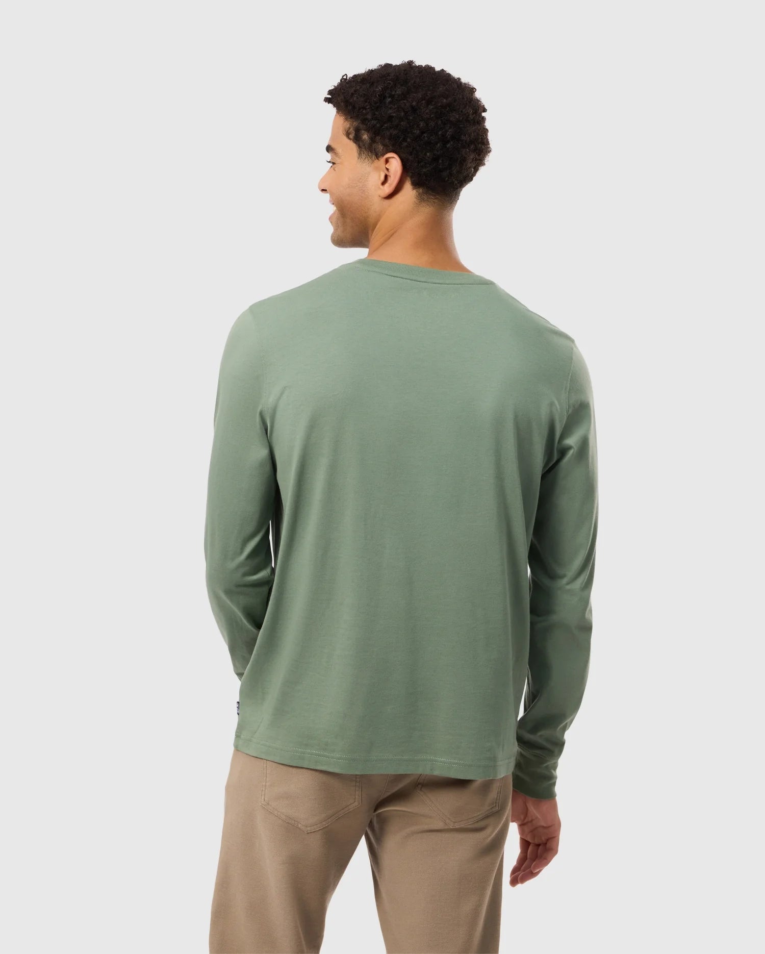 A person with short curly hair is wearing the Psycho Bunny MENS ALBERTON LONG SLEEVE TEE in light green, made from Pima cotton, along with beige pants. They are standing and facing away from the camera against a plain background.