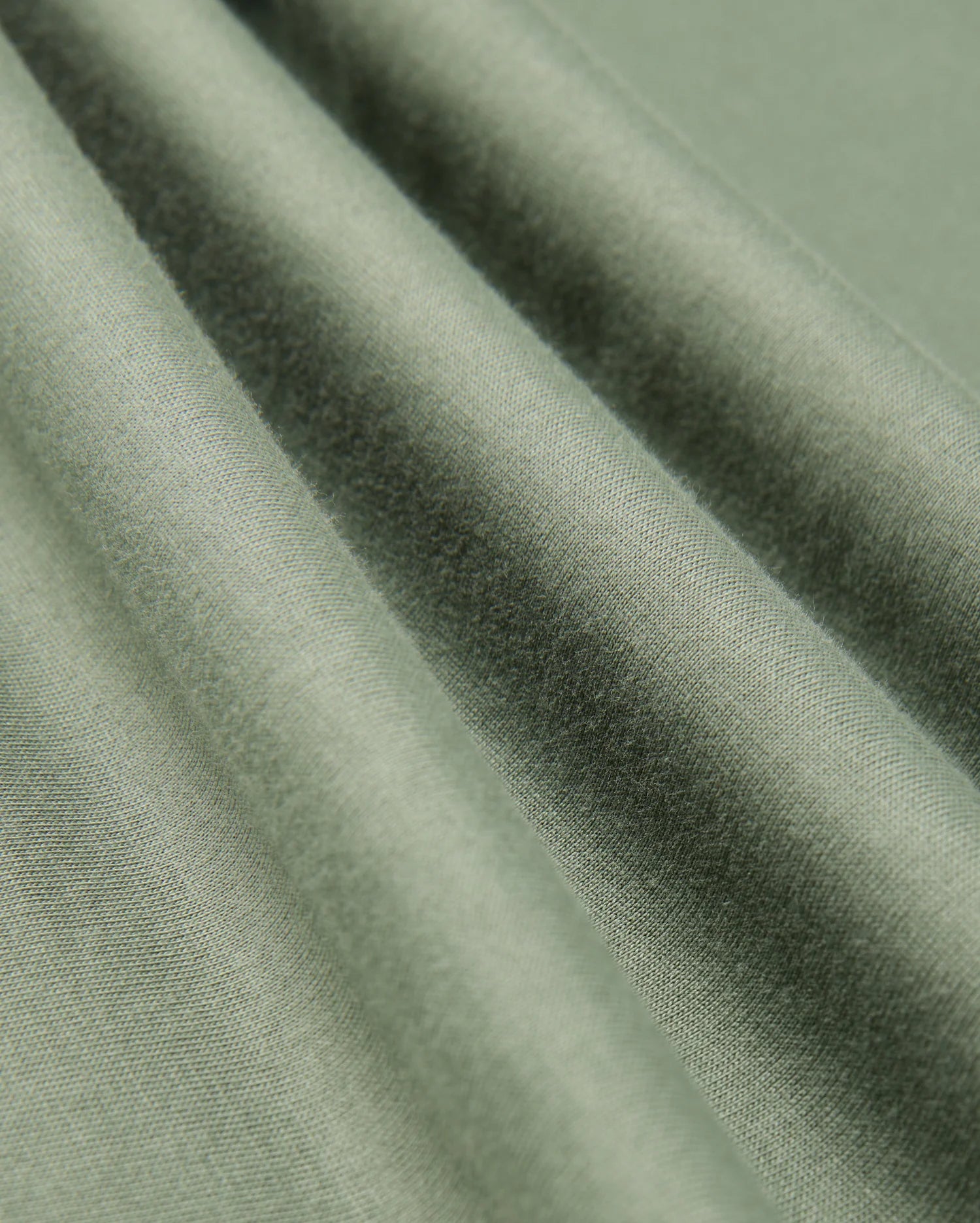 A detailed view of the Mens Alberton Long Sleeve Tee by Psycho Bunny, crafted from green Pima cotton that boasts a smooth texture and soft sheen, beautifully draped in gentle folds. The tee's weave and interaction with natural light produce delicate variations in color and shadow, elevating it to a true fashion masterpiece.